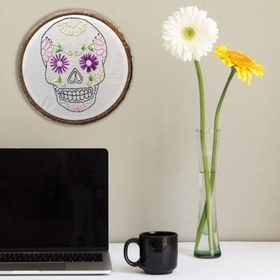 Mesa branca com portátil, caneca preta, vaso com flores e quadro redondo bordado de caveira colorida na parede