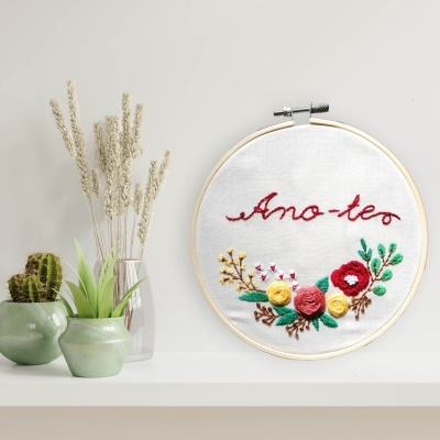 Bordado com texto 'Ano-teo' e flores coloridas num bastidor de madeira, junto a vasos com plantas.