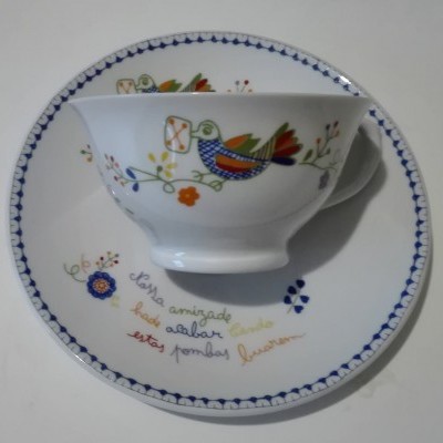 Conjunto de prato e chávena branco com padrões coloridos de pássaros e flores e texto em português.