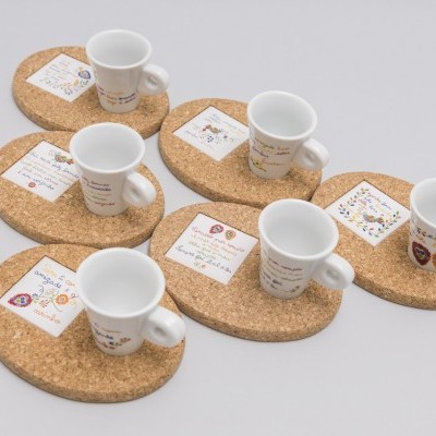 Cinco chávenas de café brancas sobre bases de cortiça com etiquetas coloridas, em fundo branco.