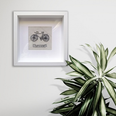 Quadro branco com ilustração de bicicleta e texto 'LET'S RIDE!' ao lado de planta verde e branca.