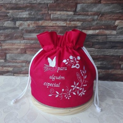 Saco vermelho bordado com cordões brancos sobre superfície clara e parede de pedra