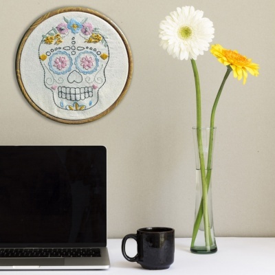 Computador portátil, caneca preta, vaso com flores branca e amarela e bordado de caveira colorida na parede