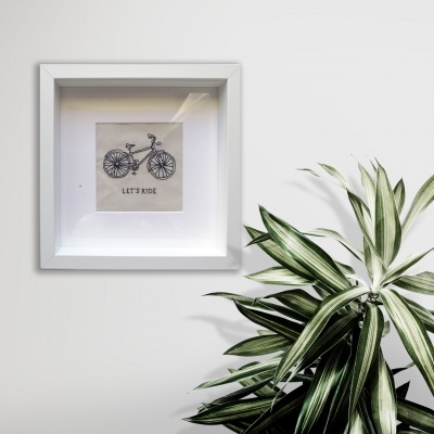 Quadro branco com ilustração de bicicleta e texto 'LET'S RIDE' ao lado de planta verde e branca