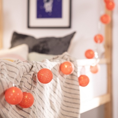 Guirlanda de luzes de bolas de tecido laranja numa cama com manta riscada branca e almofadas cinzentas