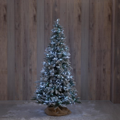 Árvore de Natal artificial com neve e luzes brancas em base coberta por tecido castanho