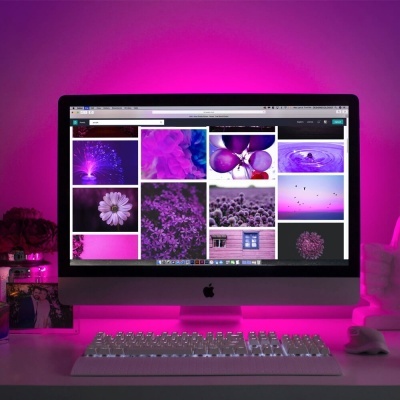 Computador Apple iMac com ecrã a mostrar imagens em tons roxo e rosa, teclado e rato brancos, luz ambiente rosa.