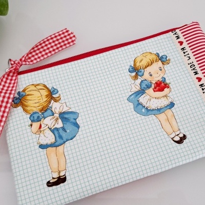 Estojo de tecido com padrão quadriculado, ilustrações de menina, faixa vermelha listrada e laço de fita