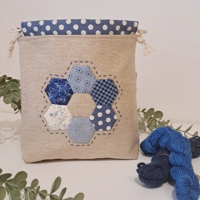 Bolsa de tecido bege com padrão de hexágonos em tons de azul e branco, decorada com ramos verdes e novelos de lã azul e cinzenta.