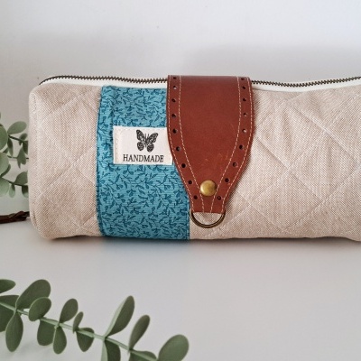 Estojo retangular bege com faixa azul, tira de couro castanho, etiqueta handmade e folha verde ao lado