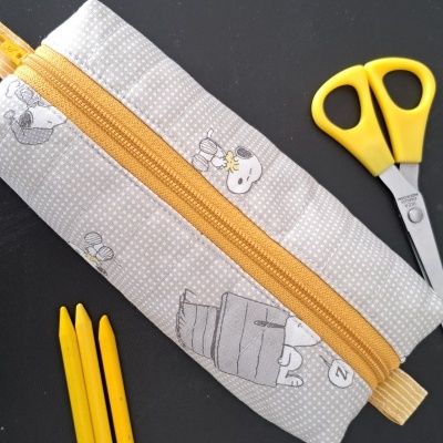 Estojo cinza claro com zíper amarelo, tesoura e lápis amarelos sobre fundo preto