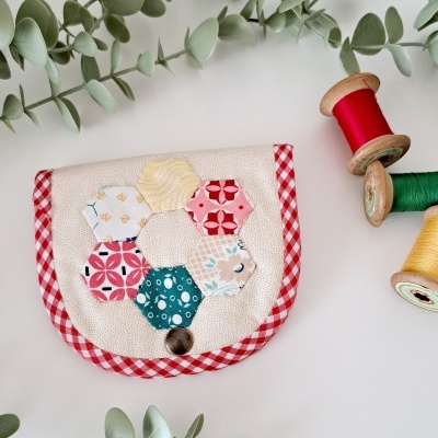 Carteira de tecido com padrão hexagonal colorido, borda vermelha quadriculada e botão metálico. Raminhos verdes e carretéis de linha colorida ao lado.