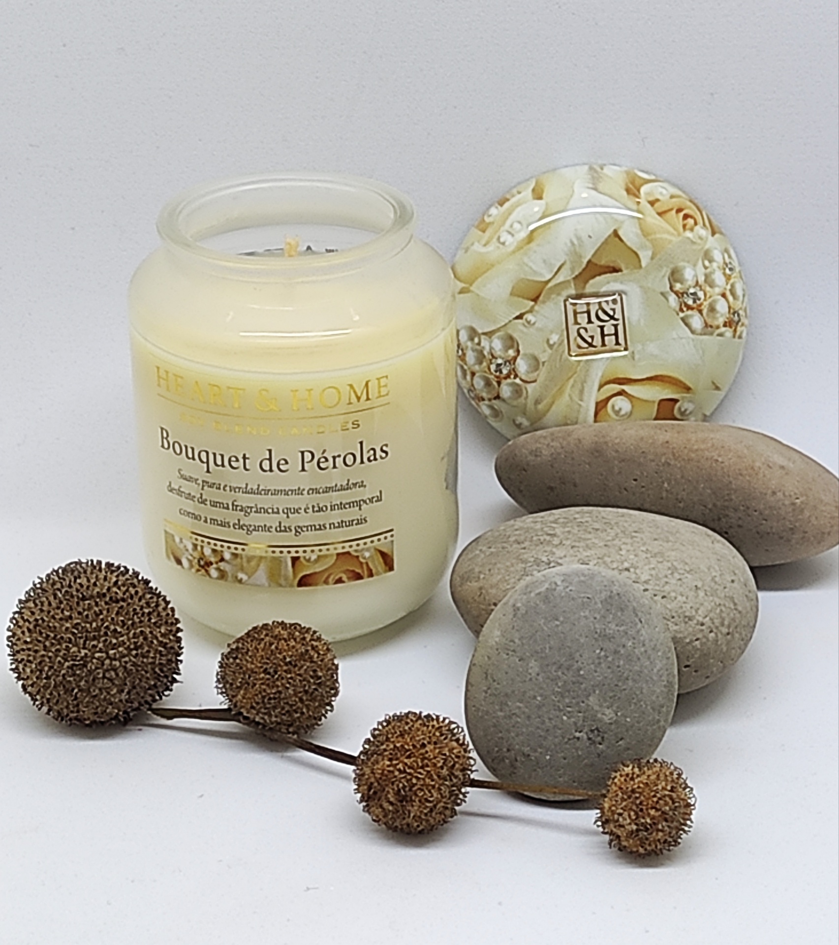 Vela perfumada em frasco de vidro branco com tampa decorada, rodeada por pedras e ramos secos