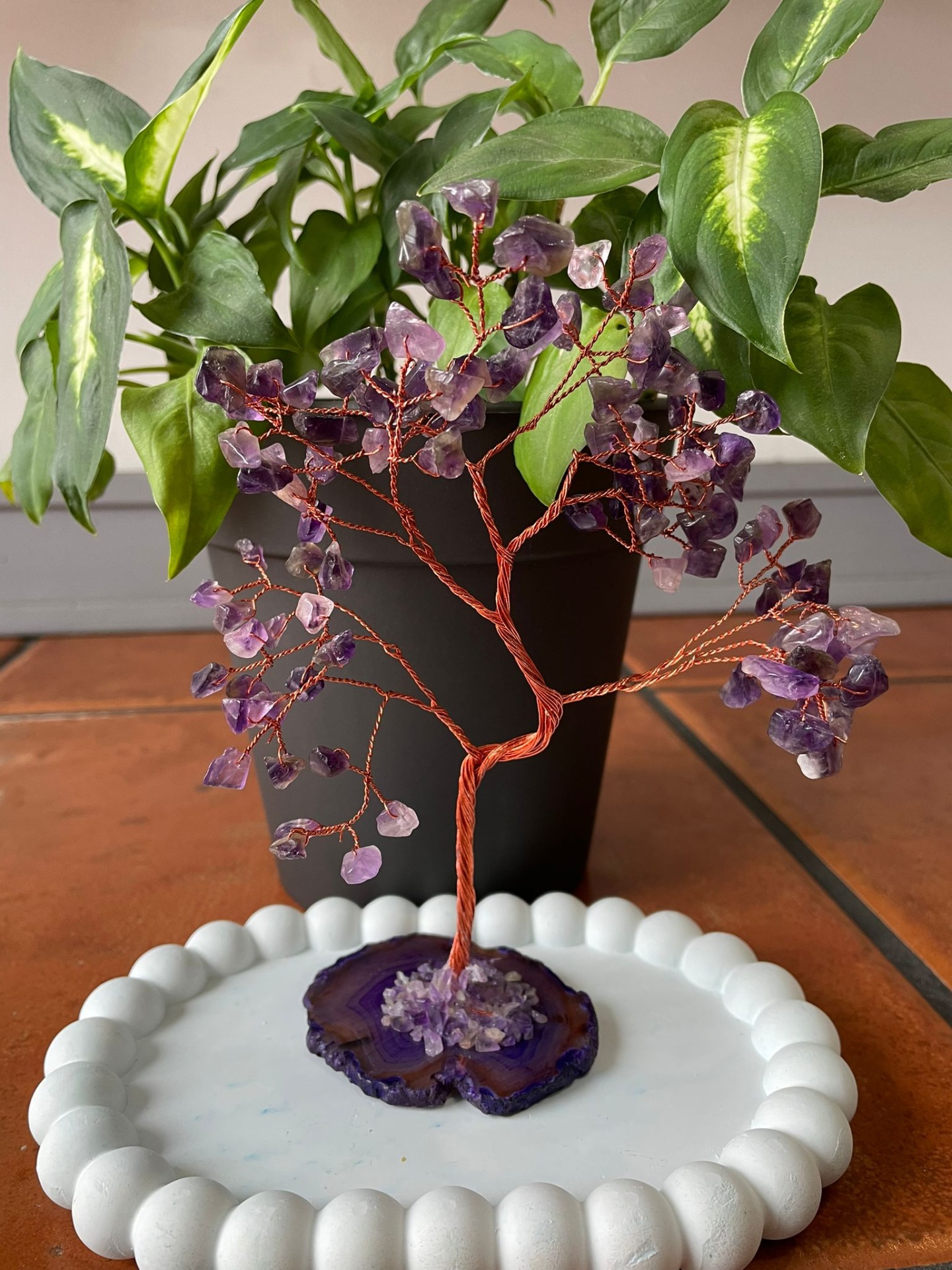 Árvore de fio de cobre com pedras roxas numa base branca, com planta verde em vaso preto ao fundo