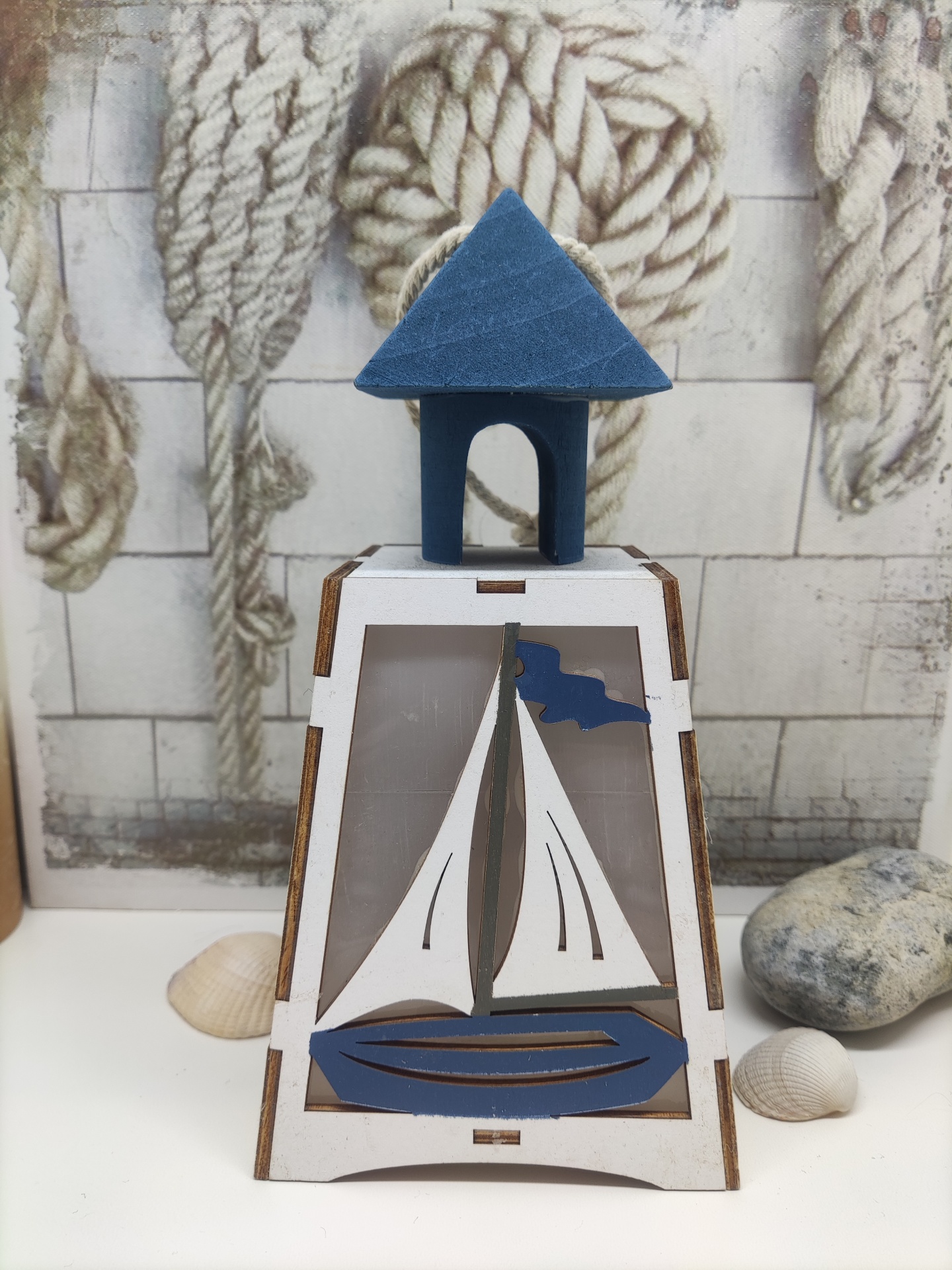 Lanterna decorativa de madeira com barco e casa azul em fundo com cordas e pedras