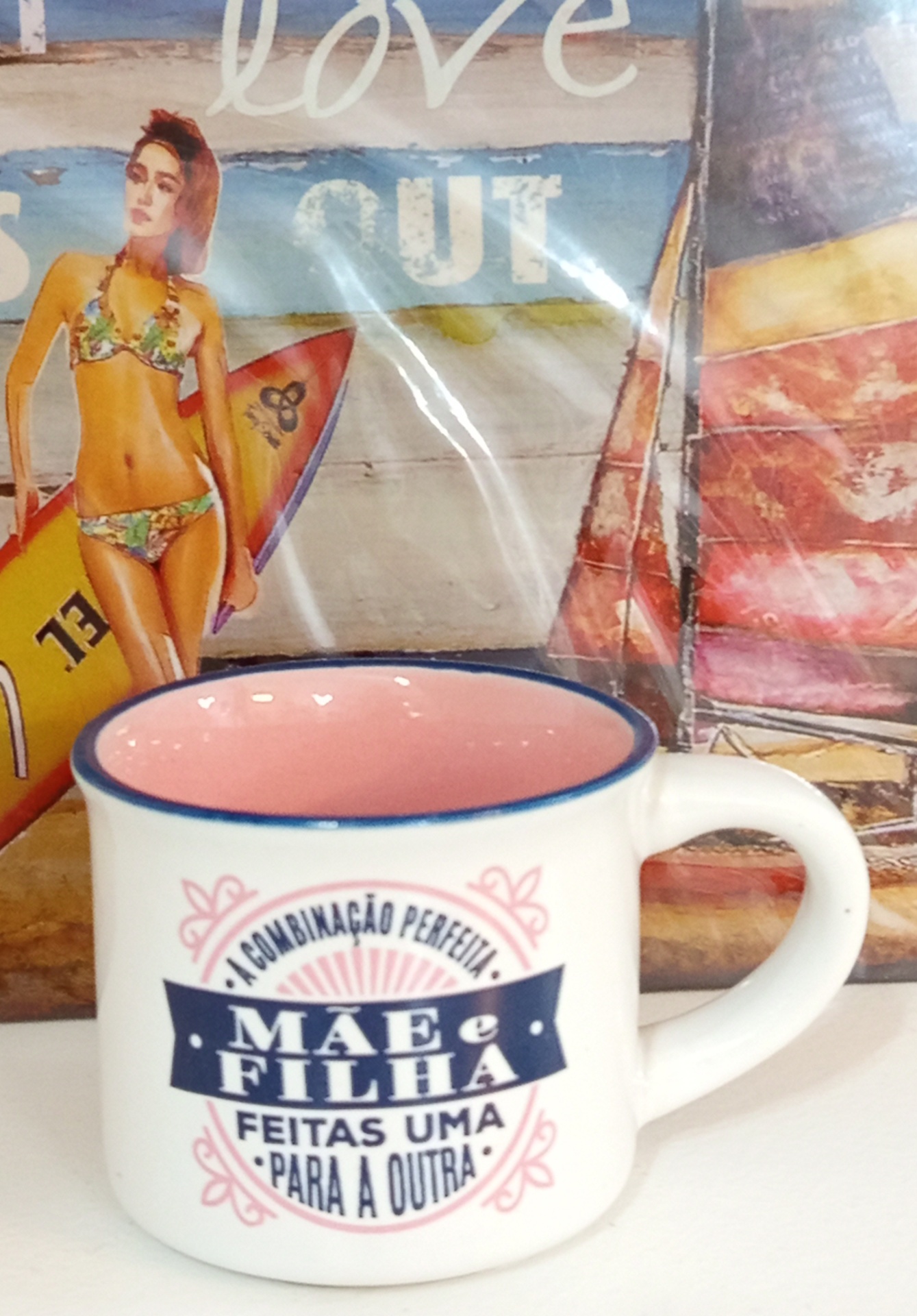 Caneca branca com interior rosa e texto em português sobre mãe e filha, em frente a imagem de mulher com prancha de surf