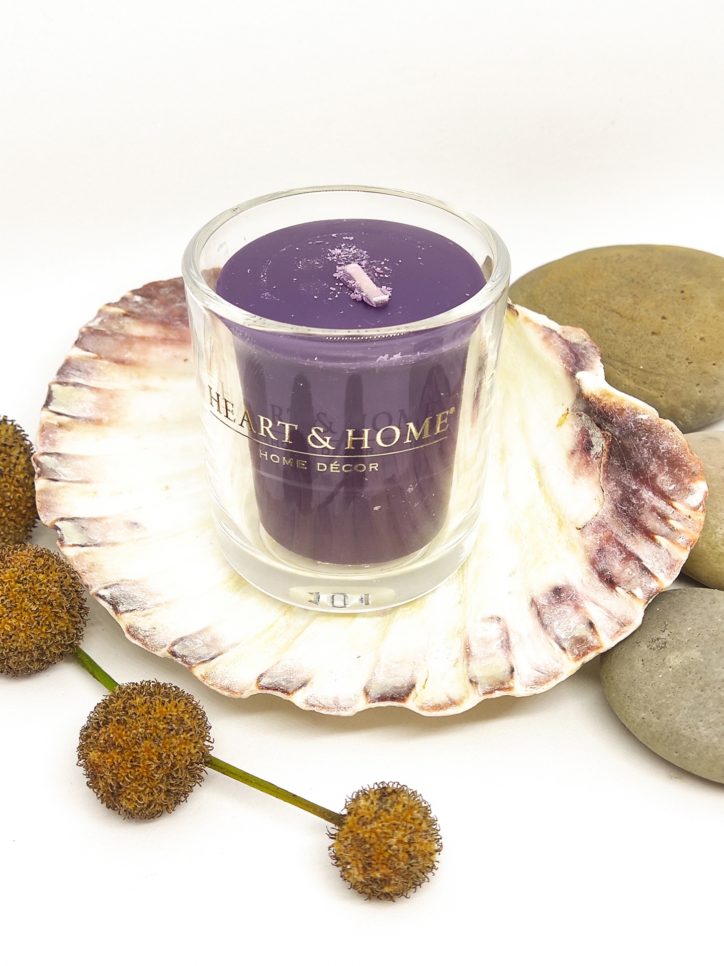 Vela roxa em copo de vidro sobre concha grande com flores secas e pedras