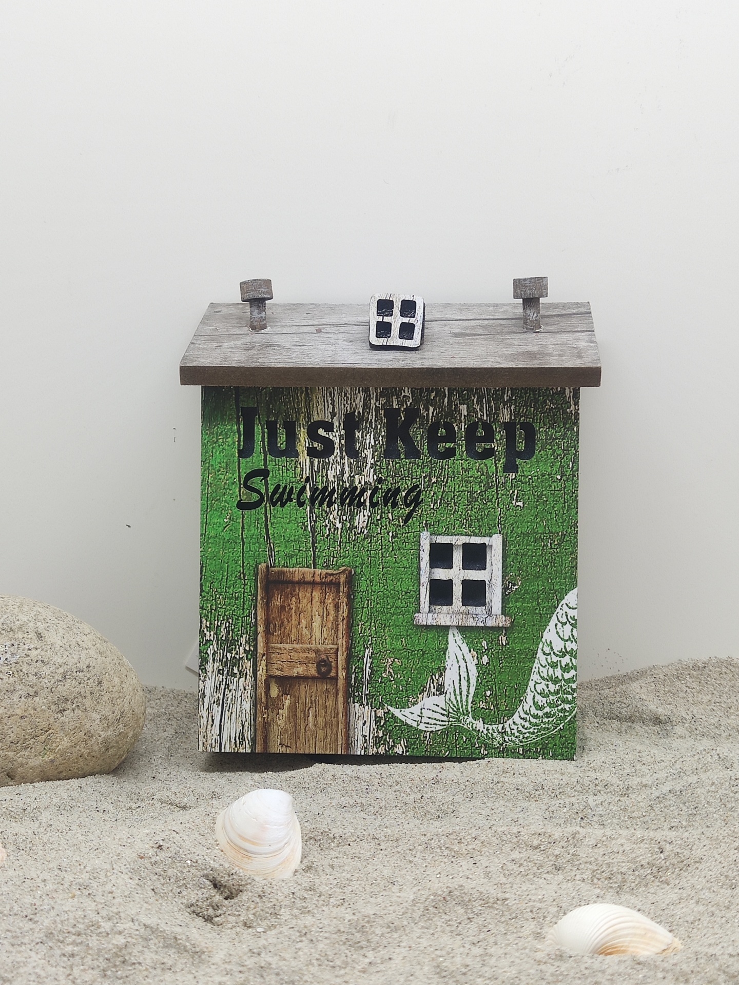 Caixa decorativa em forma de casa verde com texto 'Just Keep Swimming' sobre areia com conchas