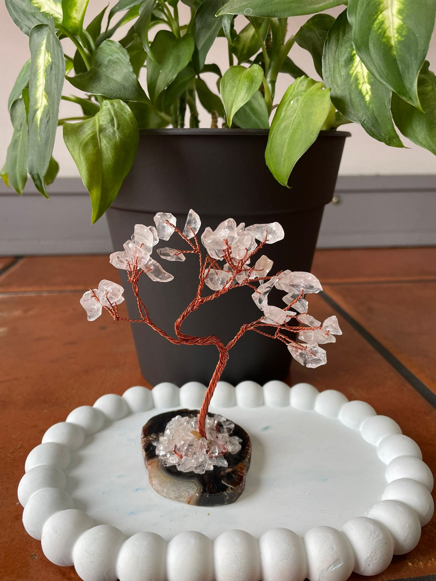 Árvore decorativa com cristais em base de pedra sobre superfície branca, vaso preto e planta ao fundo.