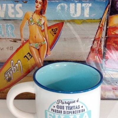 Caneca branca com interior azul e texto em português, ao lado de imagem de mulher de fato de banho com prancha de surf.