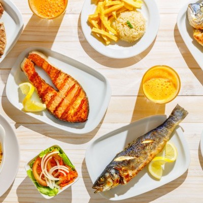 Refeições variadas em travessas brancas com peixe grelhado, arroz, batatas fritas, salada e sumo laranja sobre mesa clara.