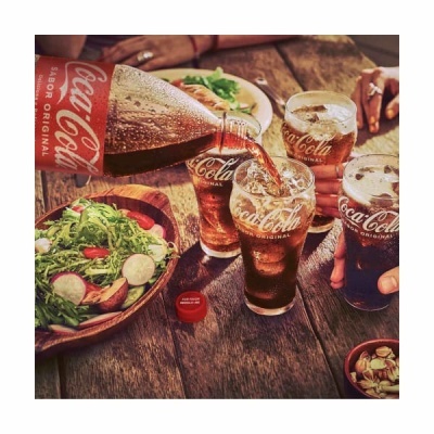 Garrafa de Coca-Cola a servir em copos com gelo numa mesa de madeira com salada e amendoins