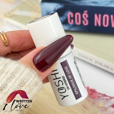 Frasco de cosmético YOSHI UV HIBRID branco com rótulo roxo, unhas vinho escuro e livro ao fundo