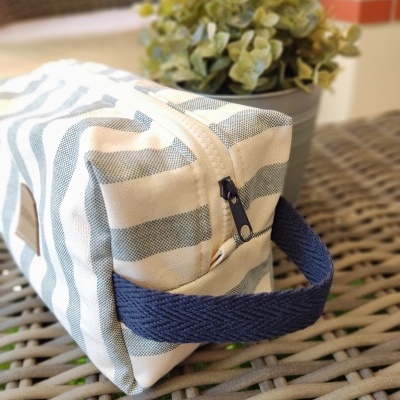 Bolsa riscada branca e cinza com pega azul e fecho preto sobre mesa de vime com planta ao fundo