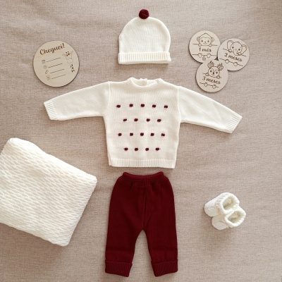 Conjunto de roupa de bebé em malha, com camisola branca e pontos bordô, calças bordô, gorro branco, meias brancas, manta e placas de madeira com texto e animais.