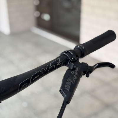 Punho e travão preto de bicicleta com cabos numa área com chão claro e parede de tijolos.