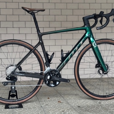 Bicicleta de estrada Scott Addict 20 M54 carbono verde e preto com pneus castanhos em suporte, fundo de parede de azulejos cinza