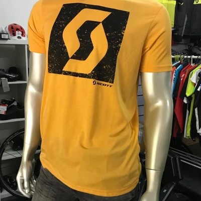 T-shirt laranja com grande logótipo preto SCOTT em manequim metálico, loja desportiva ao fundo