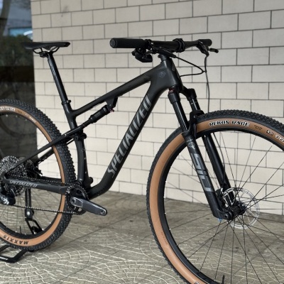 Bicicleta de montanha preta com pneus castanhos em piso com padrão circular e parede de tijolos.