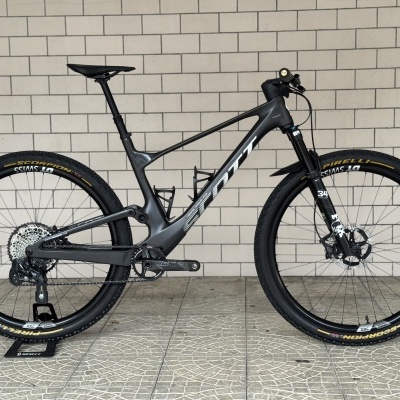 Bicicleta de montanha preta Scott com pneus Pirelli, suspensão Fox 34 contra parede de azulejos.