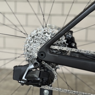 Roda traseira de bicicleta com cassete e desviador preto e cinza em fundo de parede de tijolos.