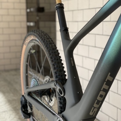 Quadro de bicicleta de montanha cinzento com detalhes dourados, roda traseira e pneu castanho claro numa parede de azulejos brancos