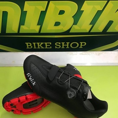 Sapatilhas de ciclismo pretas com sola vermelha e logo 'fizik' em frente a placa verde e amarela.