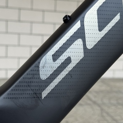 Quadro de bicicleta preto com letras brancas e textura em pontos com parafuso preto, parede de tijolos brancos ao fundo