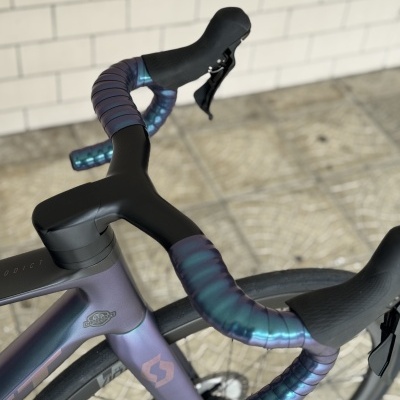 Bicicleta de estrada cinza-azulada com guiador enfaixado em fita azul iridescente e manípulos pretos junto a parede de azulejos brancos