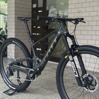 Bicicleta de montanha cinza escuro em carbono com pneus Maxxis e suspensão RockShox.