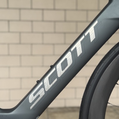 Quadro de bicicleta cinza com letra SILCOMTT em branco e roda preta junto a parede de tijolos