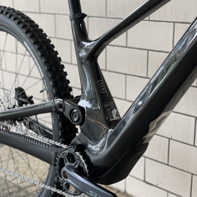 Bicicleta de montanha preta com suspensão traseira e pedaleira SLX junto a parede de azulejos cinza.