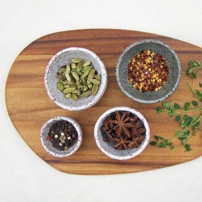 Quatro tigelas de cerâmica com especiarias em tábua de madeira oval, ramos verdes ao lado.