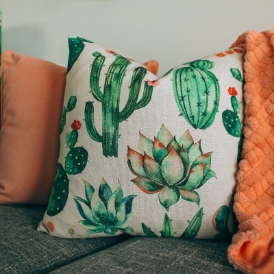 Sofá cinzento com almofadas decorativas em verde, laranja e estampado de cactos, e manta laranja macia