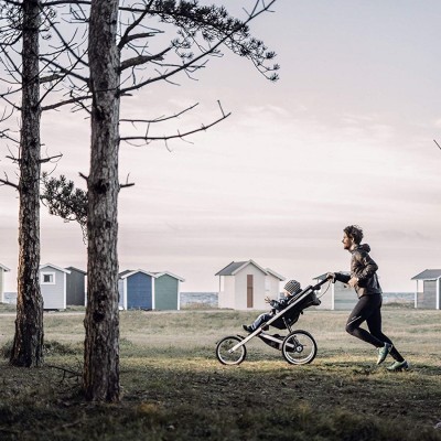 Mulher a correr com carrinho de bebé preto em campo com casas de praia ao fundo