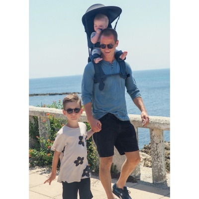 Homem com bebé num carrinho de transporte atrás e criança ao lado, junto ao mar