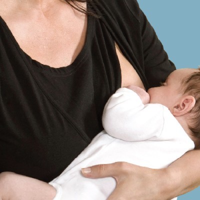 Mulher a amamentar bebé com camisola preta e macacão branco, fundo azul claro.