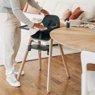 Cadeira alta para bebés em madeira clara e plástico preto a ser limpa numa sala de estar.