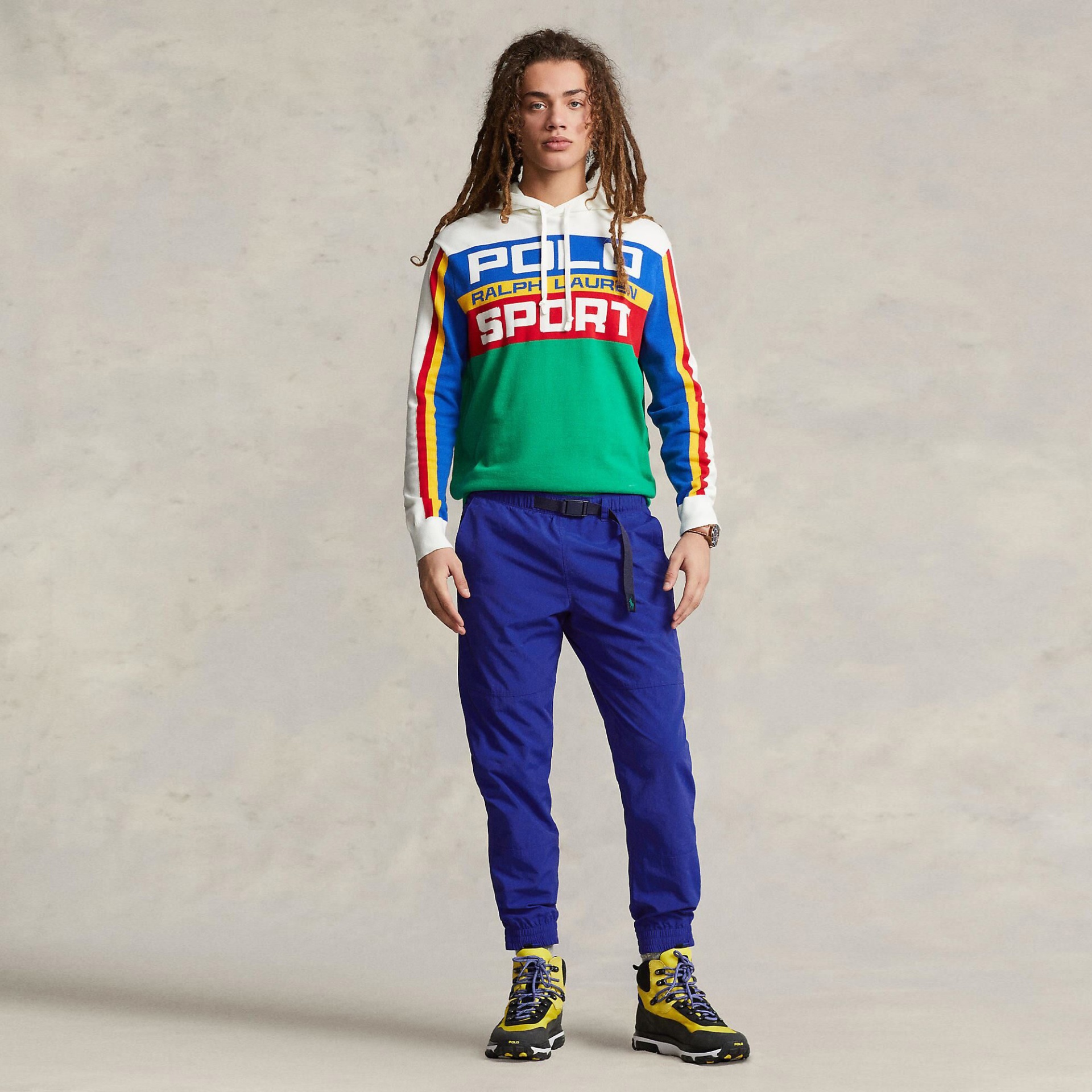 Modelo com hoodie multicolorido, calças azuis e botas amarelas em fundo claro