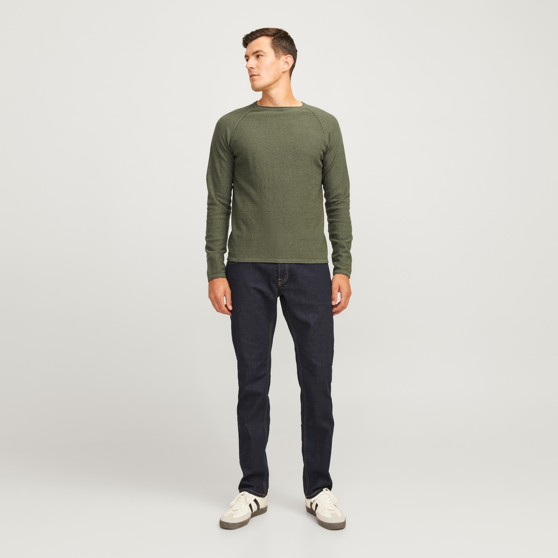 Homem com camisola verde, jeans escuro e ténis brancos num fundo branco